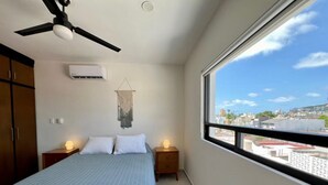 A bedroom with a bed, bedside tables, a ceiling fan, an air conditioner, and a view of the city through a large window.