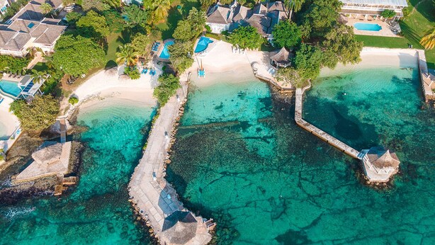 Vista aerea di un resort con spiaggia, acque turchesi cristalline e diverse strutture con tetti di paglia.