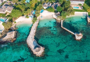 An aerial view of a resort with a beach, clear turquoise waters, and several thatched-roof structures.