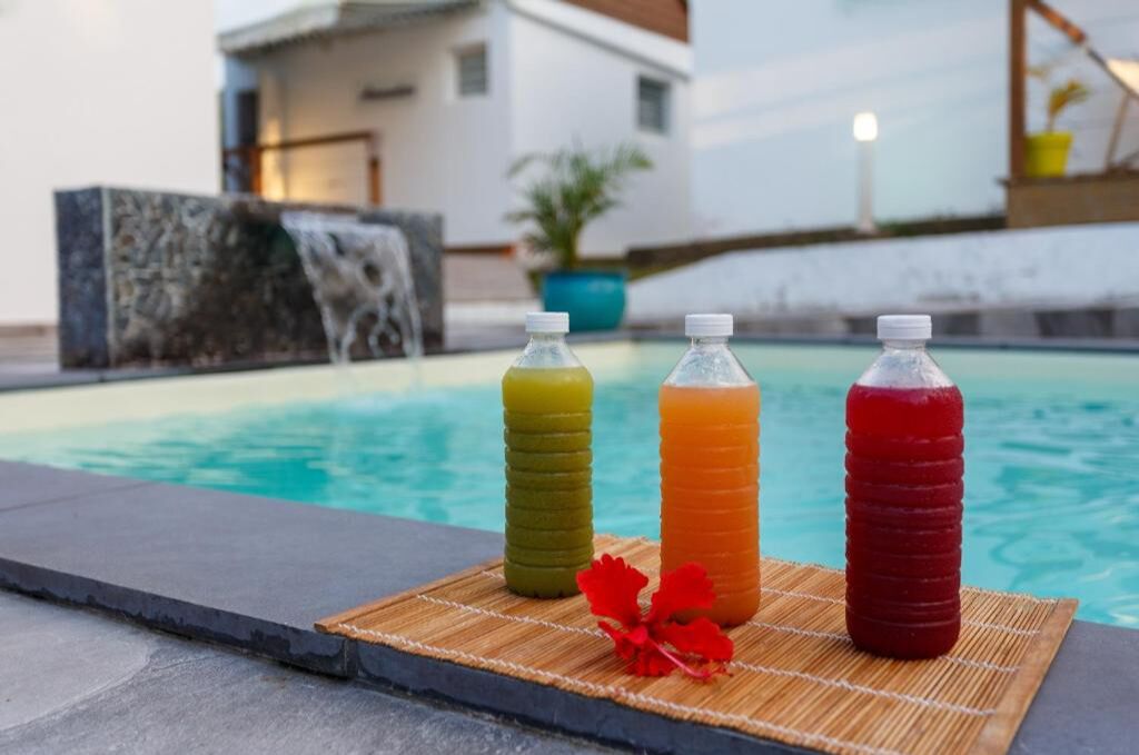 Three bottles of beverages on a mat by a pool with a house and a waterfall in the background.