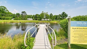 A wooden bridge leading to a body of water with a sign indicating 'Millville by the Sea' and 'Kayak & Canoe Pond'.