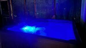 Un jacuzzi con luz azul, rodeado por una estructura de madera y una ventana con vista a árboles.