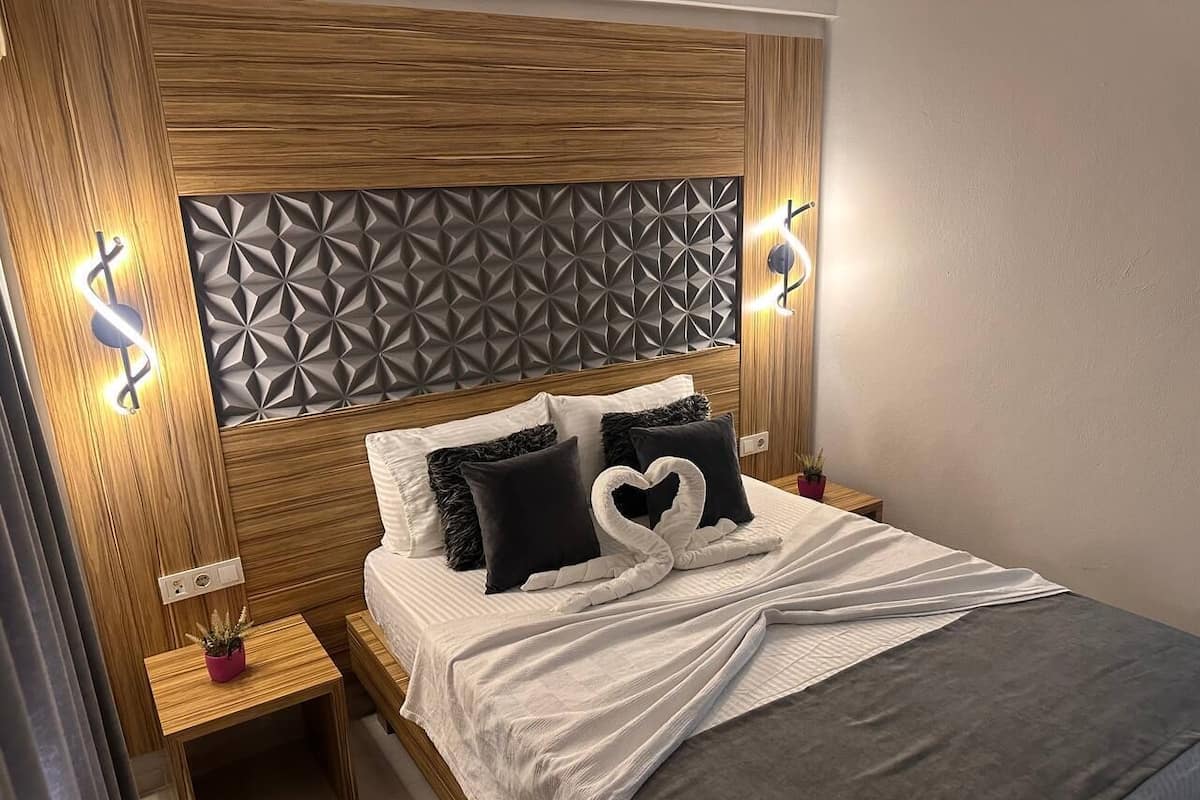 A bedroom with a wooden headboard, a bed with white bedding and black pillows, a heart-shaped towel, and a small nightstand with a plant.