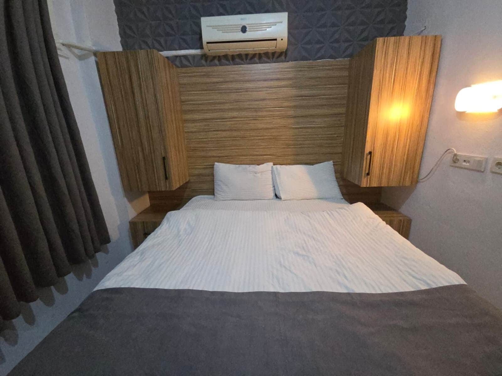 Una habitación de hotel con una cama, cabecero de madera, dos mesitas de noche, aire acondicionado de pared y una lámpara.