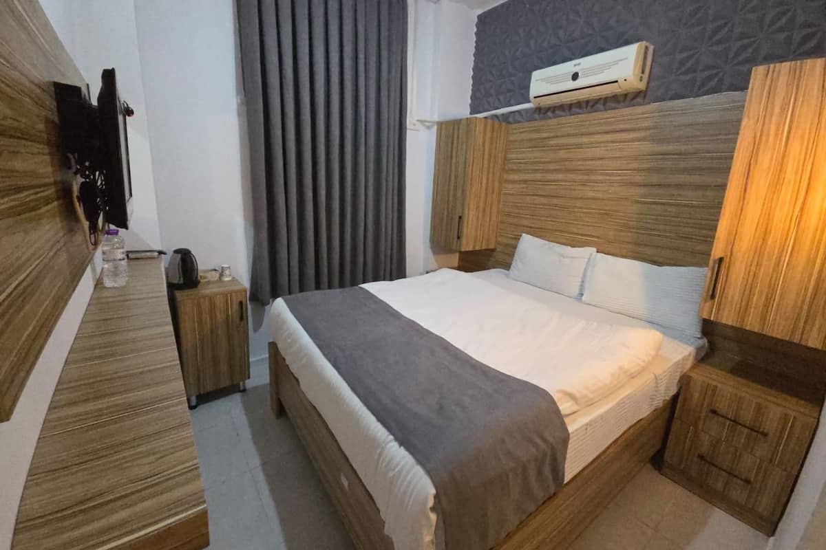 A hotel room with a wooden bed frame, a bedside table, a nightstand, a wall-mounted TV, and a window with curtains.