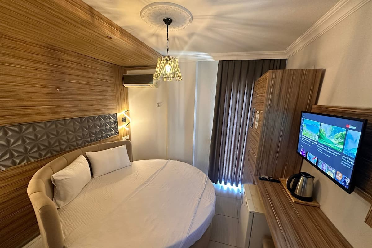 A modern hotel room with a large bed, wooden paneling, a ceiling light fixture, a television on the wall, and a window with curtains.