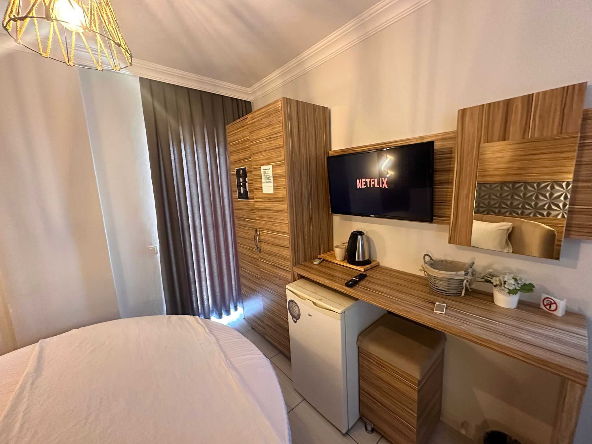 A hotel room with a bed, a small refrigerator, a coffee maker, a TV displaying 'NETFLIX', and a wooden desk with a cup and a remote control.