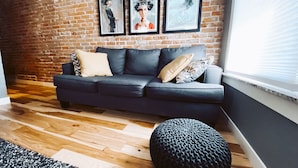 A modern living room with a dark grey sofa, a black ottoman, and three framed artworks on a brick wall.