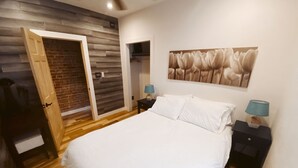 A bedroom with a wooden floor, a bed with white bedding, two bedside tables with lamps, a ceiling fan, and a brick wall.