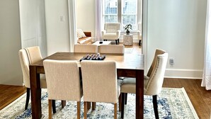 Een eetgedeelte met een houten tafel en zes stoelen, een geweven lamp en uitzicht op een woonkamer met een bank en fauteuils door een doorgang.