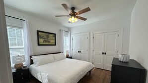 Een slaapkamer met een bed, een plafondventilator, een ingelijst schilderij en een raam met jaloezieën.