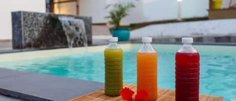 Three bottles of beverages on a mat by a pool with a house and a waterfall in the background.