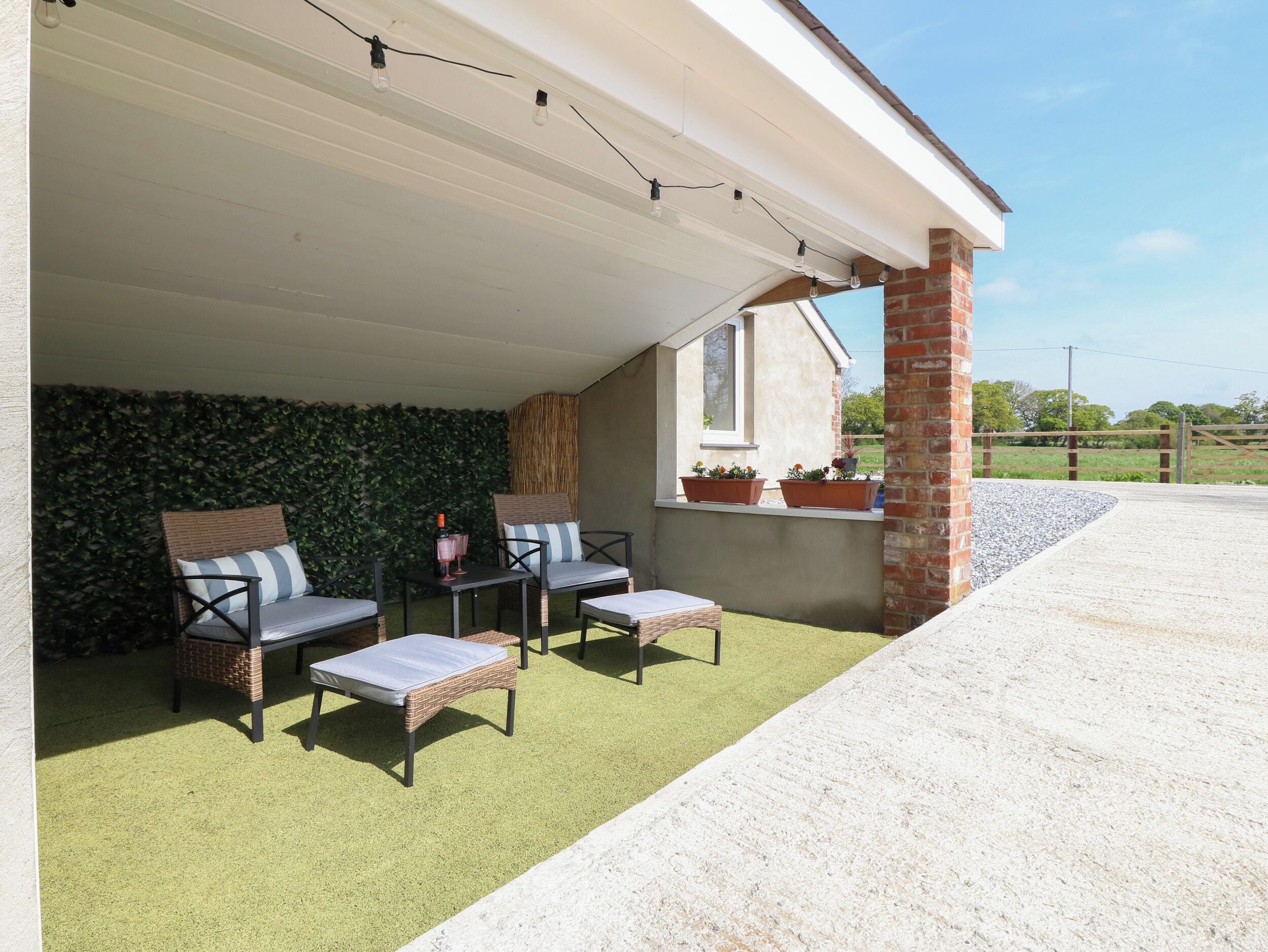 A covered outdoor seating area with wicker furniture, a green artificial turf, and potted plants on a brick wall.