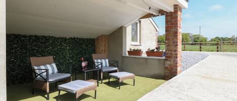 A covered outdoor seating area with wicker furniture, a green artificial turf, and potted plants on a brick wall.
