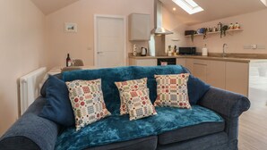A cozy living room with a blue sofa and patterned cushions, a kitchen with modern appliances, and a skylight above. - Y Felin (Caernarfon)