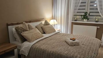 A neatly made bed with a beige blanket and pillows, a bedside table with a lamp, and a window with sheer curtains.
