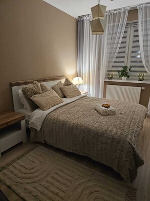 A neatly made bed with a beige blanket and pillows, a bedside table with a lamp, and a window with sheer curtains.