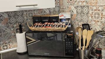 A kitchen with a microwave, tea and coffee packets, a basket of utensils, and a pot on the counter.