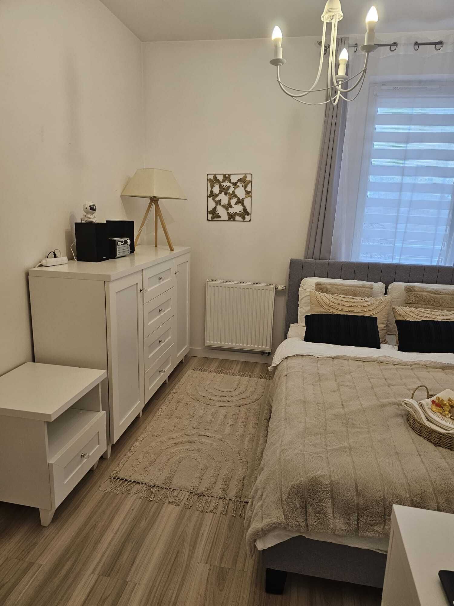 Een slaapkamer met een bed, nachtkastjes, een kaptafel, een radiator, een kroonluchter en een raam met gordijnen.