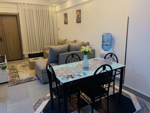 Un comedor con mesa de vidrio, sillas negras y un jarrón con flores. Hay un sofá con cojines y un dispensador de agua en la pared.