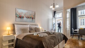 A modern bedroom with a large bed, bedside tables with lamps, a cityscape painting, and a view of the outdoors.