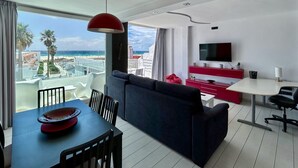 Una habitación de hotel moderna con comedor, sofá, televisor y balcón con vistas a la playa y palmeras.