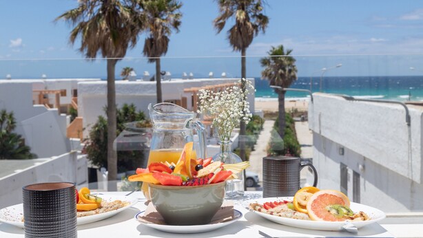 Una mesa en el balcón con un frutero, una jarra de zumo de naranja y una taza de café, con vistas a una playa con palmeras y cielo despejado.
