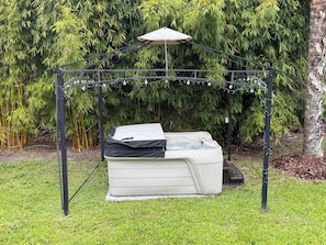 Un bain à remous installé sous une pergola en métal, illuminé de guirlandes lumineuses, et entouré de verdure.