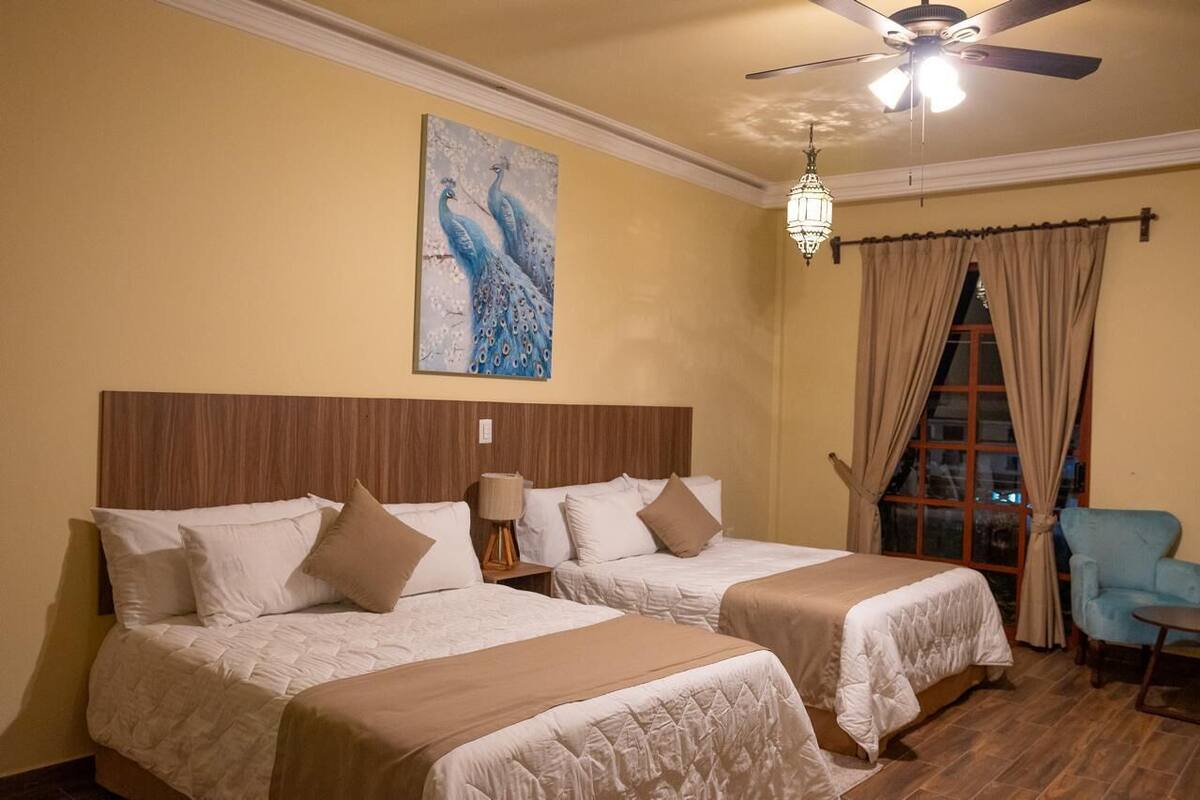 A hotel room with two beds, a peacock painting, a ceiling fan, and a window with curtains.