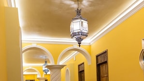 Un couloir aux murs jaunes, doté d’un sol à motifs et surmonté d’un lustre. On y trouve des colonnes, une chaise et des portes portant des numéros.