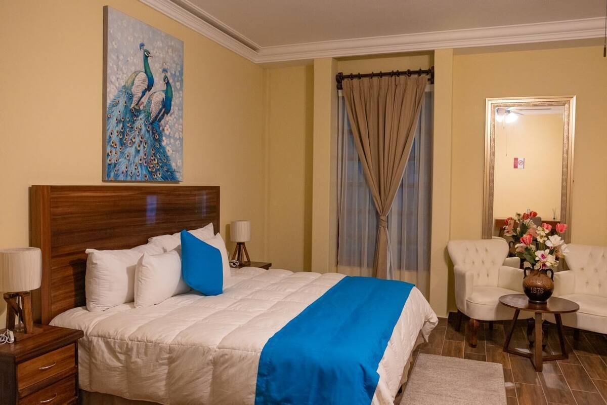 A hotel room with a large bed, a wooden headboard, a nightstand with a lamp, a chair, and a vase with flowers.