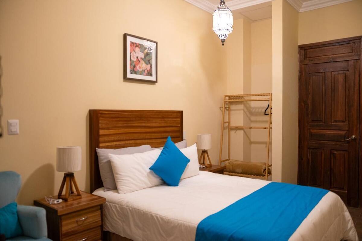 A hotel room with a wooden headboard bed, a blue throw blanket, a wooden wardrobe, and a hanging rack.