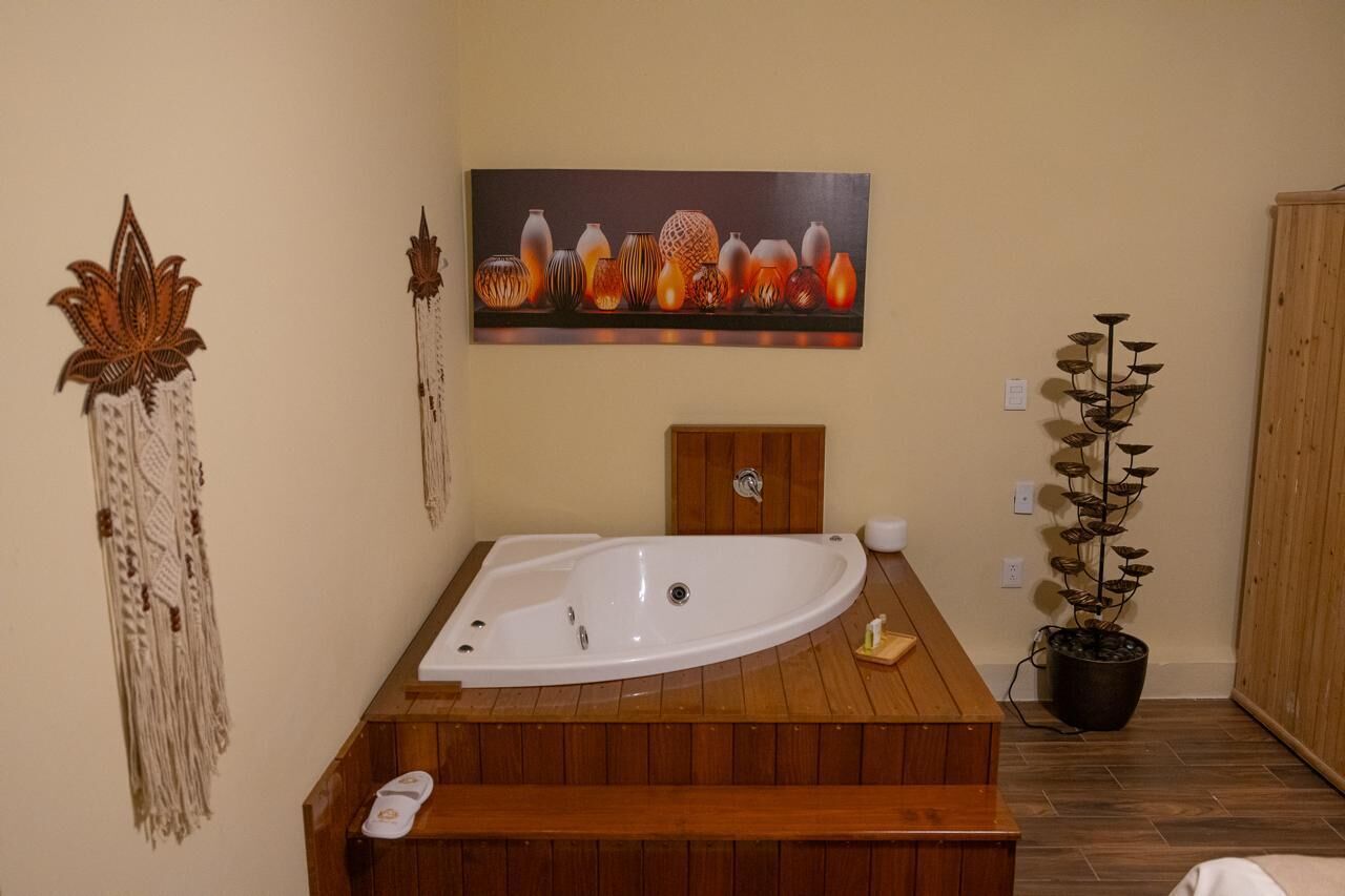 A bathroom with a wooden bathtub, a wall-mounted sink, a decorative plant, and a painting of vases.