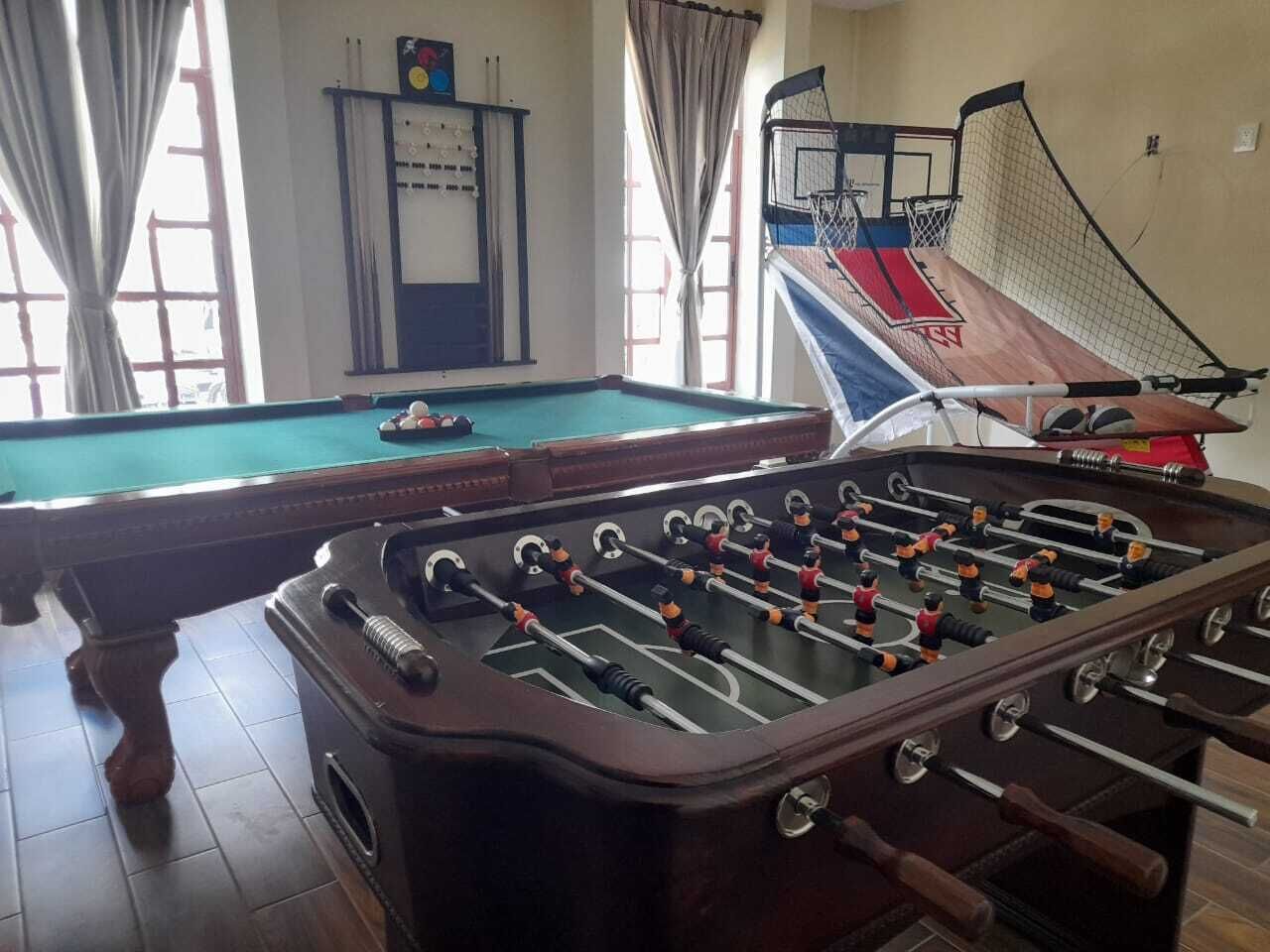 Una mesa de billar y una mesa de fútbolín en una sala con aro de baloncesto y una ventana con cortinas.