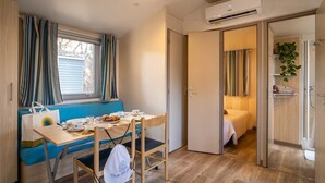 A compact room with a dining table, a bench, and a bed. There is an air conditioning unit on the wall and a plant in the corner.