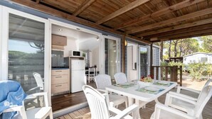 A covered outdoor dining area with a table and chairs, a kitchen visible through an open door, and a wooden deck.