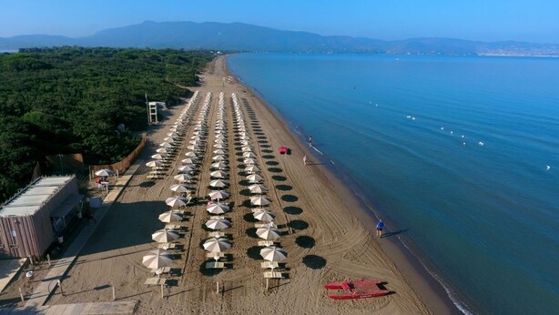 A beach with rows of umbrellas and beach chairs, a red boat, and a clear view of the sea and distant mountains.