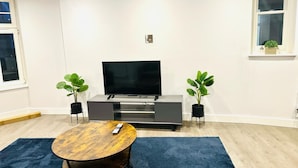 A modern living room with a flat-screen TV, a wooden coffee table, two potted plants, and a blue rug. - Spacious Margate Apartment- Beach,arts&high Street (Margate)