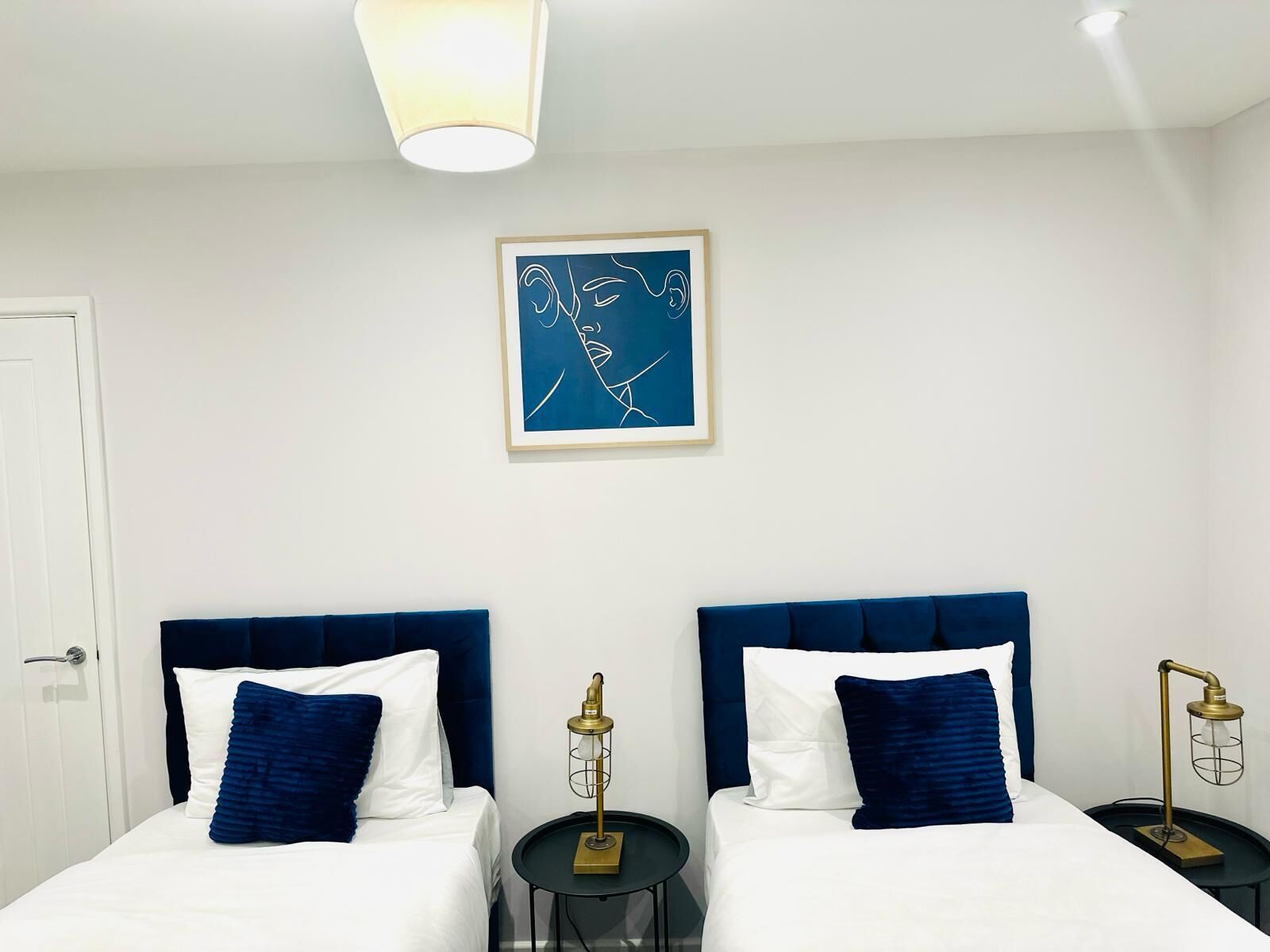 A hotel room with two beds, each with a blue headboard, white bedding, and a blue pillow. There is a framed abstract artwork on the wall above the beds.