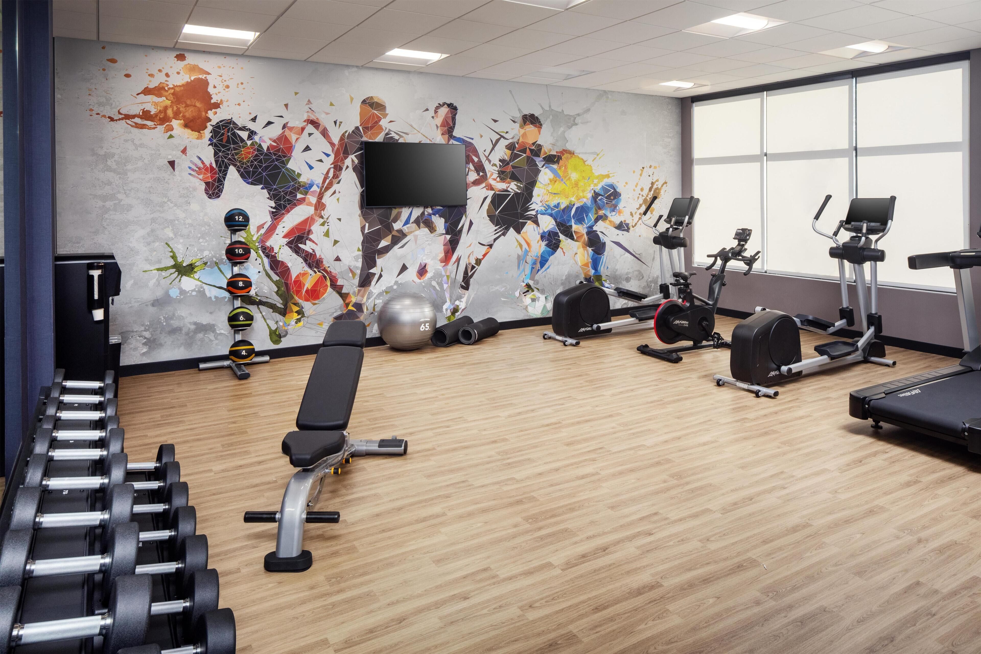A gym with exercise equipment, a wall with a dynamic mural, and large windows.