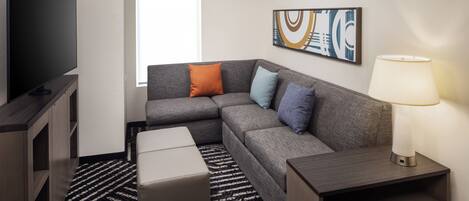A modern hotel room with a sectional sofa, a flat-screen TV, a lamp, and a painting on the wall.