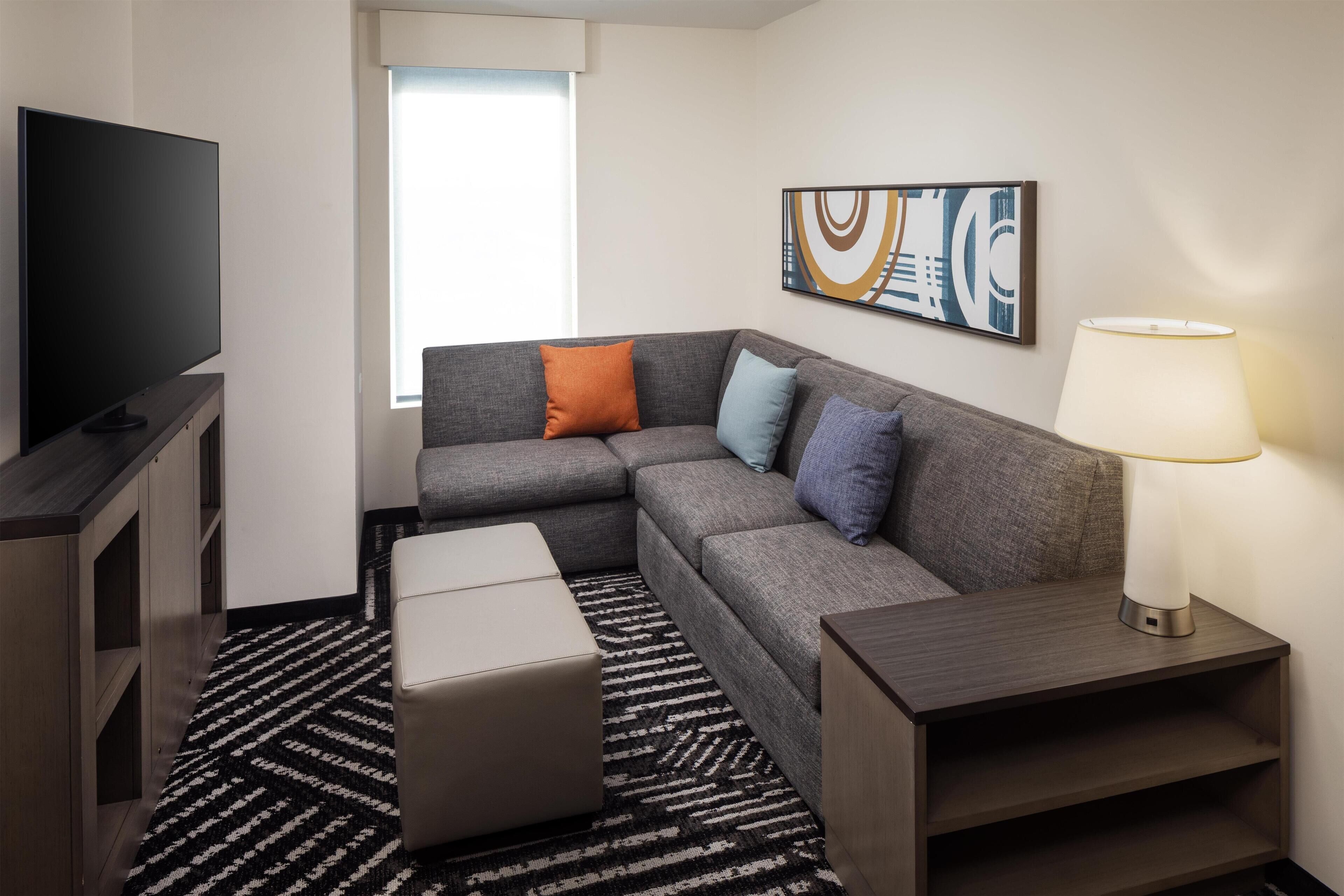 A modern hotel room with a sectional sofa, a flat-screen TV, a lamp, and a large window providing natural light.