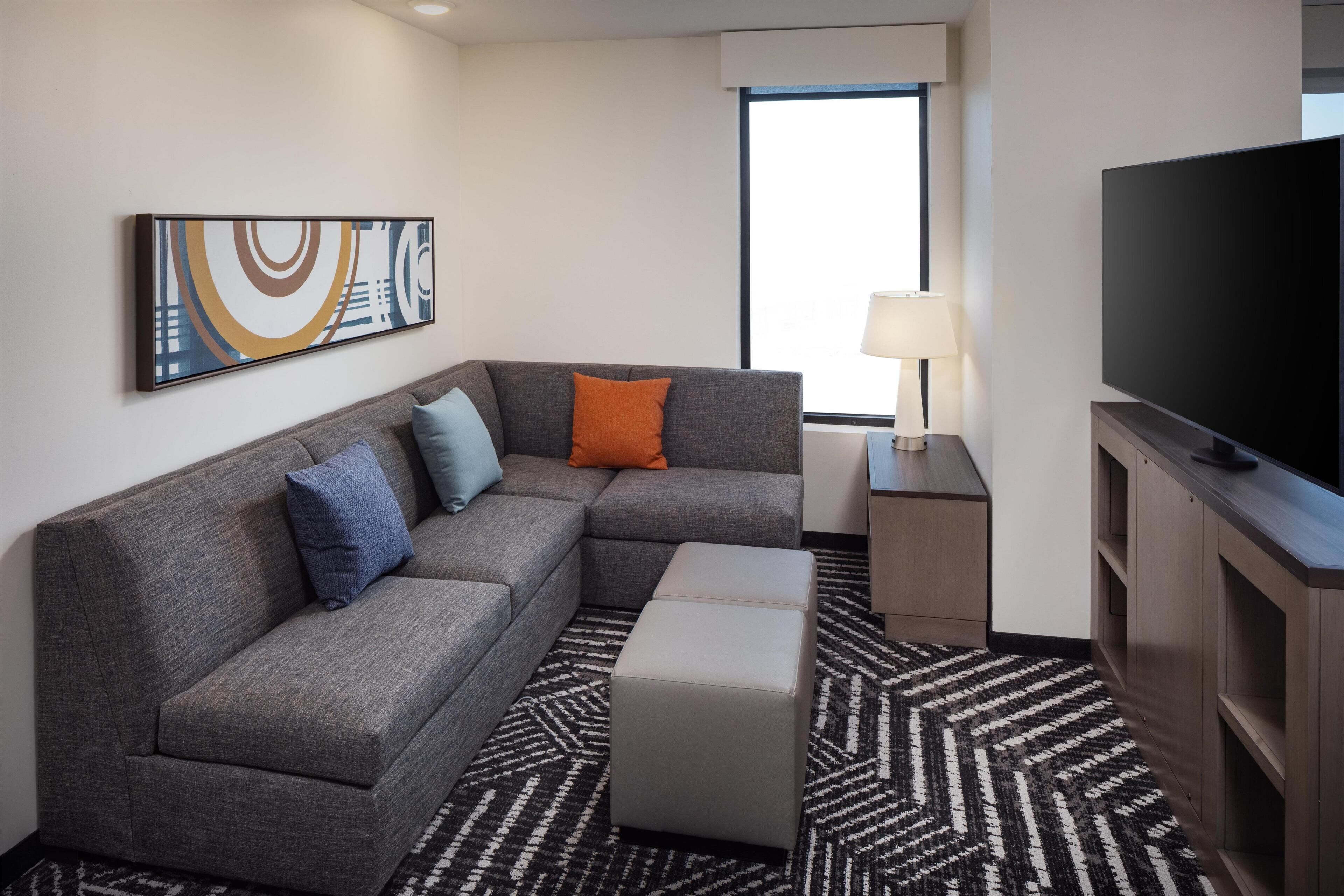 A modern hotel room with a gray sectional sofa, a flat-screen TV, a patterned rug, and a large window.
