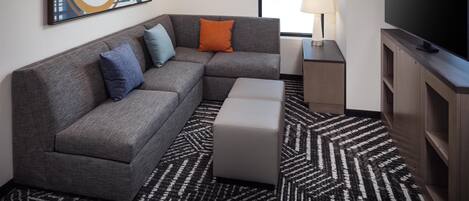 A modern hotel room with a gray sectional sofa, a flat-screen TV, a patterned rug, and a large window.