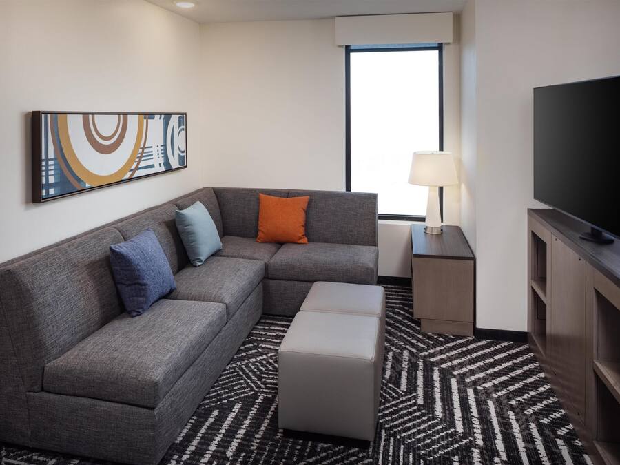 A modern hotel room with a gray sectional sofa, a flat-screen TV, a patterned rug, and a large window.