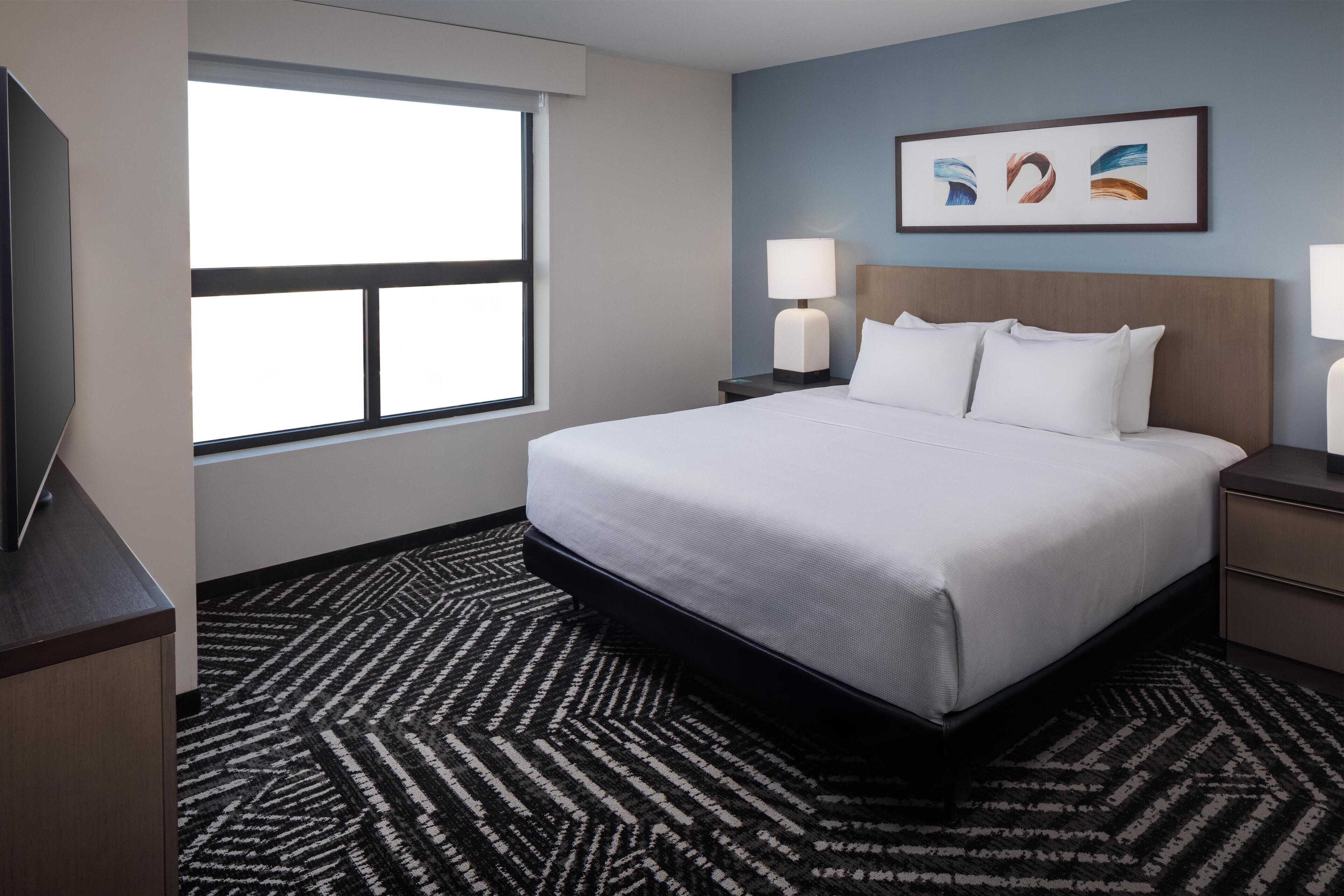 A hotel room with a large bed, two bedside tables with lamps, a flat-screen TV, a window with blinds, and a patterned carpet.