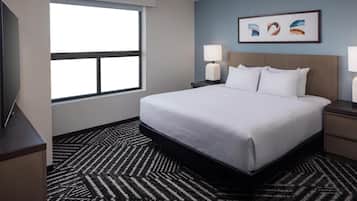 A hotel room with a large bed, two bedside tables with lamps, a flat-screen TV, a window with blinds, and a patterned carpet.
