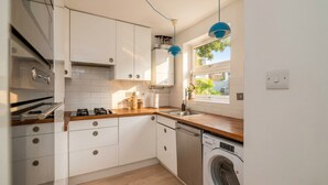 A compact kitchen with white cabinets, a dishwasher, a washing machine, and a window with a view of greenery. - Canal View House | 2BR with Garden & Parking (London)