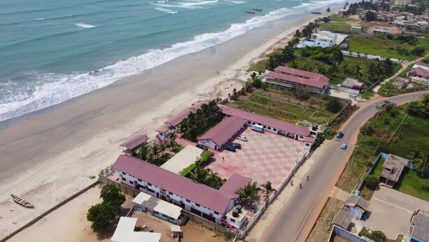 An aerial view of a coastal area with a beach, a road, and several buildings with red roofs. - Beautiful Hotel In Greater Accra Region 1 (Oshiyie)