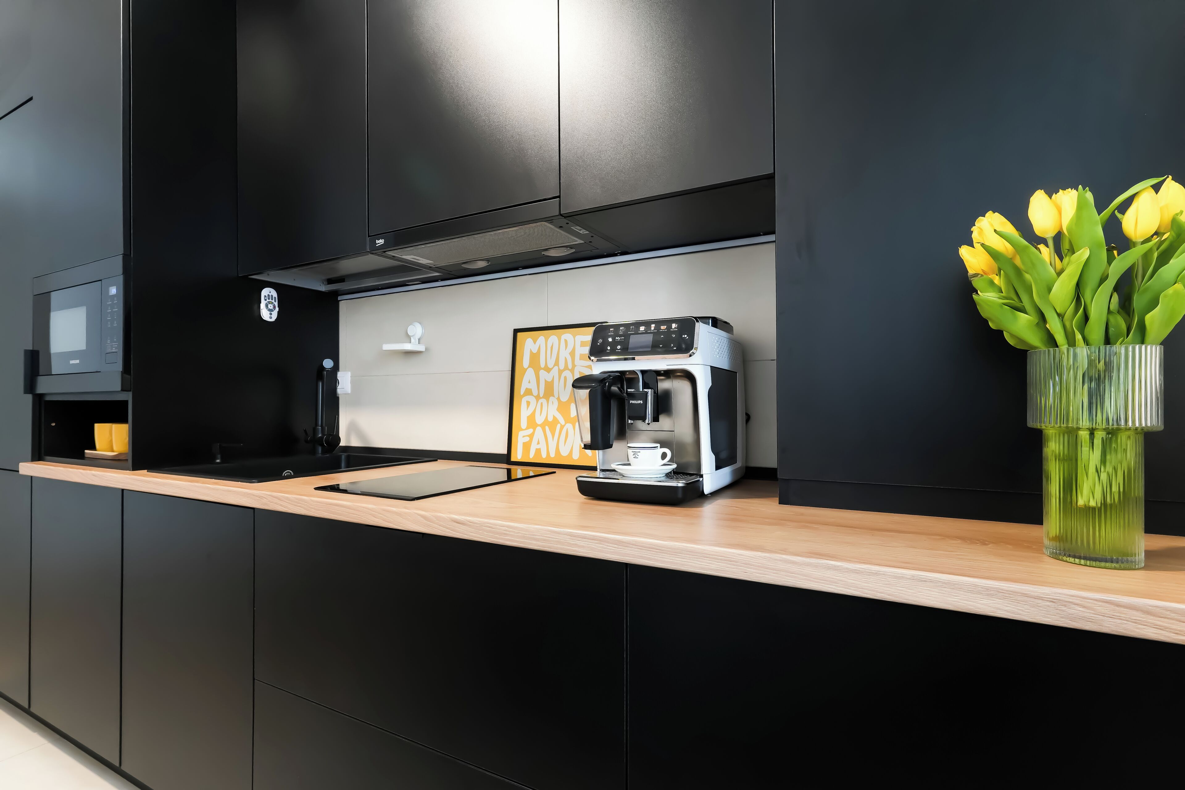 A modern kitchen with black cabinets, a wooden countertop, a coffee machine, and a vase with yellow flowers.